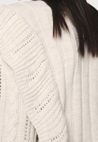 Gros plan d'une personne portant un pull en tricot de couleur crème avec des motifs en torsades et en côtes, et des cheveux longs, raides et foncés reposant sur l'épaule.