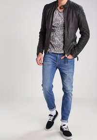 Giacca da motociclista in pelle nera con spalle trapuntate, t-shirt grigia con motivo, jeans azzurri chiari e sneakers nere con suola bianca e calzini.