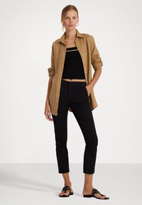Lichtbruine linnen button-up shirt over een zwarte cropped top, gecombineerd met strakke zwarte ankle-lengte broeken en zwarte sandalen met decoratieve banden.