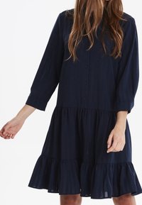 Abito blu navy in tessuto leggero, con maniche lunghe, chiusura a bottoni sul davanti e orlo a balze a strati. Una texture sottile arricchisce l'interesse visivo.