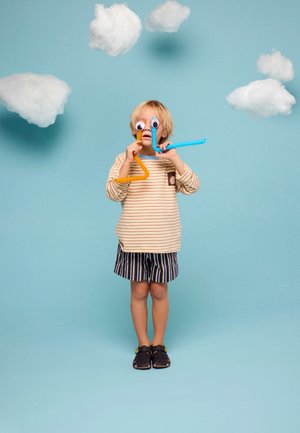 Bambino con maglietta a righe e pantaloncini che tiene in mano tubi flessibili colorati con grandi occhi di carta su uno sfondo blu con nuvole di carta.