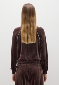 Tuta in velluto marrone con una vestibilità comoda, dotato di polsini e vita a costine, e piping a contrasto lungo le spalle. Capelli che scendono sotto le spalle.