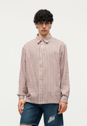 Νέος άνδρας με σγουρά μαλλιά που φορά μακρυμάνικο μπεζ και καφέ καρό πουκάμισο και σκισμένα μπλε τζιν σορτς, στέκεται μπροστά σε απλό φόντο.