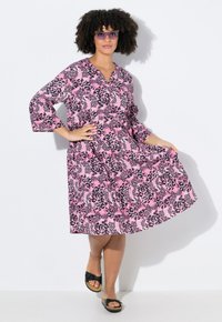 Rosa Leopardenmusterkleid mit V-Ausschnitt, 3/4-Ärmeln und lockerer Passform. Mit schwarzen Akzenten und einem fließenden Rock. Schwarze Rutschen-Sandalen.