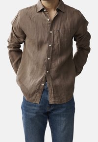 Hnědá lněná košile s dlouhým rukávem, knoflíky, náprsní kapsou a volným střihem. V kombinaci s modrými džíny. Viditelný strukturovaný povrch.
