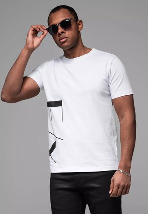 Hvid t-shirt med et sort geometrisk design på fronten, parret med mørke jeans. Model iført sorte solbriller, stående mod en grå baggrund.