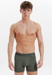 Mörkgröna boxerkalsonger med en mjuk textur, märkeskant och en figurnära design, visade på en person med en neutral bakgrund.