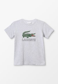 Harmaa puuvillainen lyhythihainen T-paita, jossa on vihreä krokotiililogo ja alla mustalla teksti "LACOSTE".