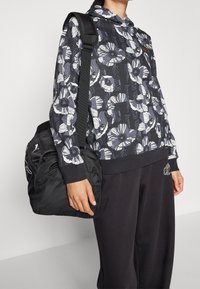 Person trägt einen schwarzen Kapuzenpulli mit Blumenmuster und schwarze Hosen, hält eine schwarze Puma-Tasche mit Schultergurt.