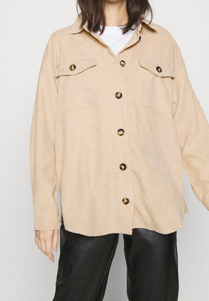 Femme portant une chemise en velours côtelé beige boutonnée sur un top blanc, associée à un pantalon noir texturé, debout devant un fond uni.