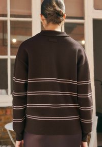 Nainen, jolla on tumma hiuskius ja joka on ponnarilla, pukeutuneena mustaan neuleeseen, jossa on valkoisia vaakaraitoja, seisoo sisätiloissa ikkunan ja tiiliseinän lähellä.