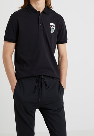 Schwarzes Polohemd mit kurzen Ärmeln, mit einem bunten Cartoon-Charakter auf der linken Brust bestickt. Wird mit schwarzen Hose mit Kordelzug kombiniert.