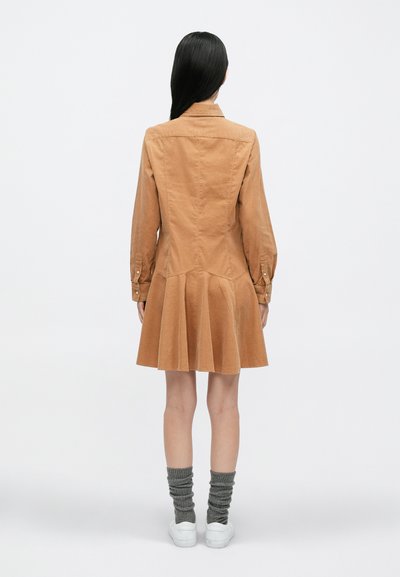 Καφέ φόρεμα από κορντούρο με εφαρμοστό μπούστο και φουσκωτή φούστα. Διαθέτει μακριά μανίκια και μανσέτες με κουμπιά. Φορεμένο με γκρι κάλτσες και λευκά αθλητικά παπούτσια.