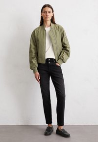 Olivgrüne Bomberjacke mit gerippten Bündchen und Saum, kombiniert mit schwarzen Slim-Fit-Jeans und schwarzen Loafers, vor einem schlichten Hintergrund.