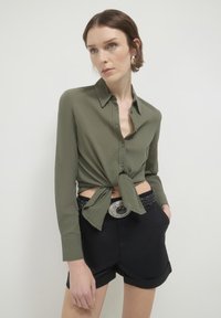 Camicia a maniche lunghe color verde oliva con nodo frontale e chiusura a bottoni, abbinata a pantaloncini neri e una cintura con fibbia argentata. Tessuto liscio.