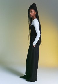 Fekete, testhez simuló felső, hosszú fehér ujjal, laza fekete nadrággal és fekete cipővel párosítva. Haj hosszan font zsinórban. Egyszerű háttér gradiens.