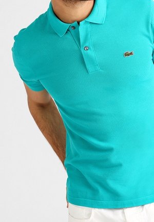 Hombre que lleva una camiseta polo de manga corta turquesa con un pequeño logo de cocodrilo en el pecho y pantalones blancos sobre un fondo liso.