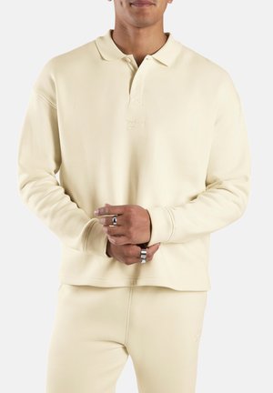 Hombre con polo de manga larga Reebok color blanco roto y pantalones a juego, con anillos plateados en los dedos, frente a un fondo liso.