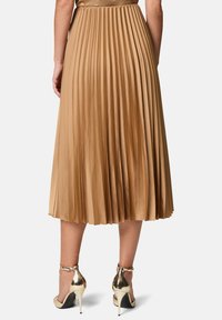 Plissé midi-rok van lichtbruin satijn met een aansluitende taille, vloeiende silhouet en glanzende gouden hoge hakken met enkelbandjes.