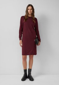 Frau steht in einem knielangen, burgunderfarbenen Kleid mit langen Ärmeln, schwarzen Stiefeletten und hält eine kleine schwarze Clutch.