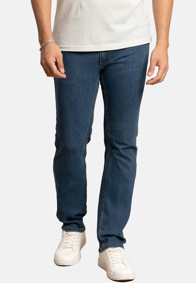 Jeans de mezclilla oscura con un corte slim, que presentan detalles de costura y una cintura media. Combinados con zapatillas bajas blancas.
