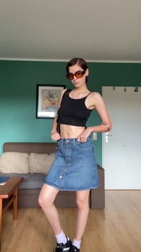 Gonna di jeans con bottoni frontali, crop top nero scuro e occhiali da sole marroni. Tessuto liscio, vestibilità casual, abbinata a scarpe sportive e calze bianche.
