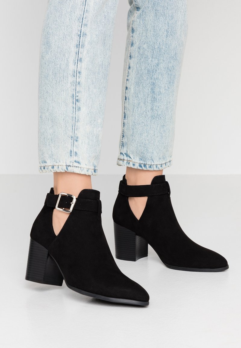 Svarta mocka ankelboots med klack och sidbuckle-remmar, bärs med ljusblå cropped jeans på en slät golv.