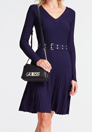 Vrouw draagt een marineblauwe geribbelde jurk met lange mouwen en bijpassende riem, en houdt een zwarte GUESS schoudertas met een gouden kettingband vast.