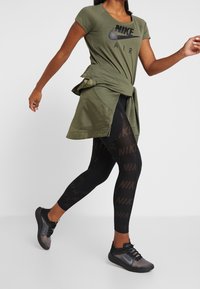 Top de Nike de color verde oliva con mangas cortas y cintura anudada, combinado con leggings negros que tienen un patrón repetido del logo de Nike y zapatillas negras.