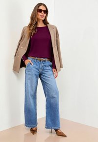 Femme portant des lunettes de soleil, un blazer à carreaux marron drapé sur les épaules, un haut violet, un jean bleu à jambes larges, une ceinture marron et des escarpins marron pointus.