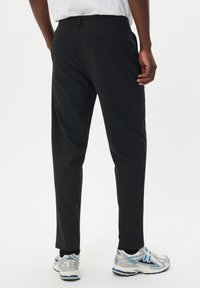 Pantaloni neri con una texture liscia, vestibilità slim e due tasche posteriori. Indossati con scarpe da ginnastica grigie e blu.