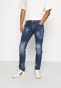 Vybledlé modré džíny s opotřebovanými detaily, slim fit, pět kapes a kontrastní švy. Nošeny s bílými teniskami.