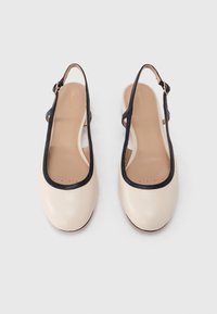 Chaussures slingback beige avec un bord noir. Tige en cuir lisse, bout arrondi, et bride ajustable avec une boucle dorée. Semelle intérieure beige clair.
