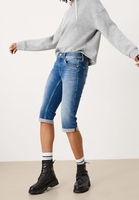 Džínové šortky v středně modré barvě, s ohrnutým lemem a designem s pěti kapsami. Kombinováno se šedou mikinou a černými šněrovacími botami.