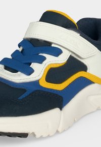 Gros plan d'une basket bleu marine avec des lacets bleus, une sangle Velcro blanche, un liseré jaune et une semelle blanche sur un fond clair.