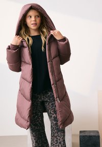 Różowy, pikowany, długi płaszcz z kapturem, zapinany na zamek błyskawiczny, z ukośnym przeszyciem. W połączeniu z czarną koszulą i rozkloszowanymi spodniami w panterkę.