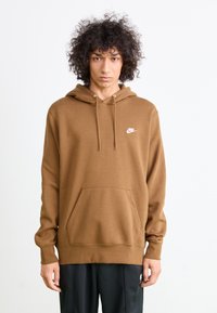 Person med krøllete hår som har på seg en brun Nike hettegenser og svarte bukser, stående mot en helt hvit bakgrunn.