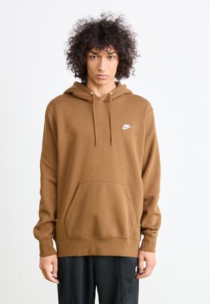 Persona con capelli ricci che indossa una felpa brown Nike e pantaloni neri, in piedi su uno sfondo bianco semplice.