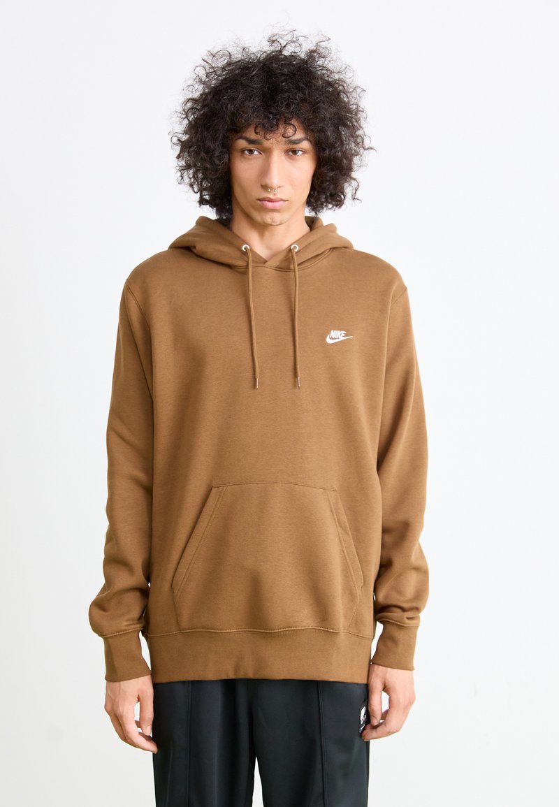 Person med krøllete hår som har på seg en brun Nike hettegenser og svarte bukser, stående mot en helt hvit bakgrunn.