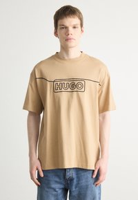 Beige bomull t-skjorte med rund hals og korte ermer. Har en svart "HUGO" logo omkranset i et rektangulært design over brystet.