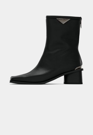 Must musta kõrge säärega must nahkne saabas terava ninaga, keskmise kõrgusega plokknõel, ja hõbedase tooni logo detail tagumise tõmbluku lähedal.