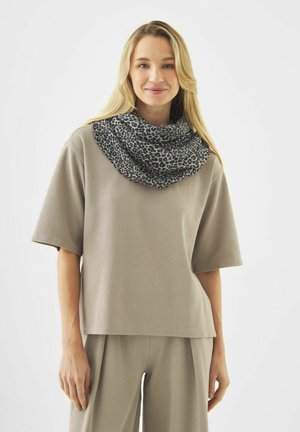 Jeune femme aux longs cheveux blonds portant un haut et un pantalon amples beige, avec un foulard léopard noir et gris autour du cou.