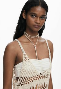 Noor naine, kellel on pikad tumedad juuksed, kannab valget kortsutatud bandeau topi ja kreemjat heegeldatud vestit, mille kaela on seotud õhuke nöörkaelakee.