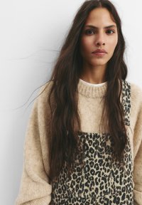 Beige gebreid sweater met een hoge hals, gedragen over een legerprint tuinbroek. De tuinbroek heeft verstelbare banden en zakken. Lang donker haar.