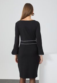 Schwarzes Kleid mit langen Ärmeln und Glockenmanschetten, mit weißen Paspeln an der Taille und den Manschetten. Glatter Stoff mit taillierter Silhouette.