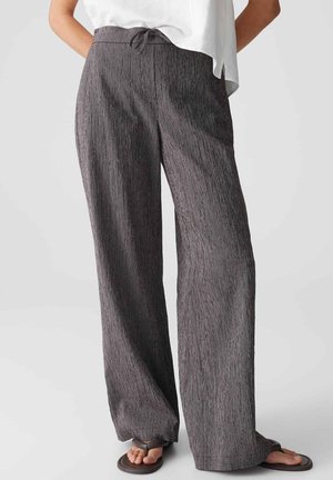 Femme portant un pantalon gris ample à cordon de serrage et des sandales noires, debout devant un fond clair uni.
