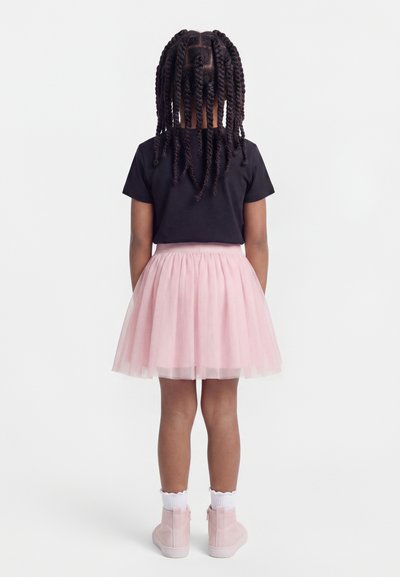 Παιδί με στριμμένα κοτσιδάκια, φορώντας μαύρο μπλουζάκι, ανοιχτό ροζ φουστίτσα από τούλι, άσπρες κάλτσες και ροζ μποτάκια αστραγάλου, στέκεται με την πλάτη.