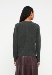 Donna con lunghi capelli ricci che indossa un maglione grigio scuro e una gonna in pelle bordeaux scuro, vista da dietro su uno sfondo bianco.