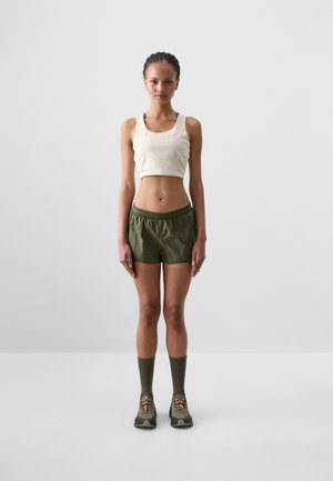 Lichtbeige crop top met dunne bandjes, gecombineerd met olijfgroene shorts. Olijfgroene sokken en sneakers maken de sportieve look af, met een minimalistisch ontwerp.