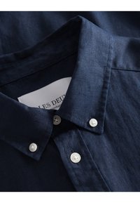 Tummansininen pellavapaita, jossa on napillinen kaulus, valkoiset napit ja teksturoitu kangas. Labelissa lukee "LES DEUX." Lähikuva yksityiskohdasta.